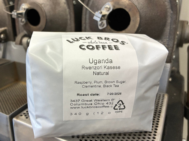 A white bag of Luck Bros. Coffee, Uganda Rwenzori Kaseke Natural roast, photographed in front of a stainless steel coffee roasting machine.
